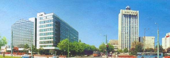 Cityscape with tall buildings, a blue sky, and a street with some trees. One building has 