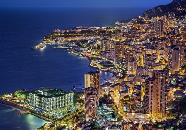 Night view of Monaco cityscape along the coast. Tall buildings and illuminated streets line the water's edge and climb the hillside.