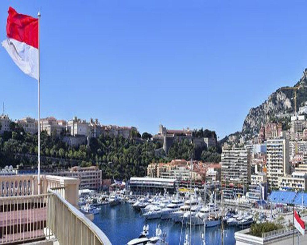 Monaco harbor scene with the Monaco flag waving. Boats fill the water, buildings line the shore, and mountains rise in the background under a blue sky.