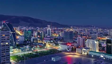 Nighttime cityscape of Ulaanbaatar, Mongolia, featuring lit skyscrapers, a central square, and mountains in the distance.