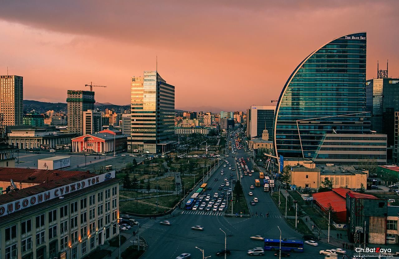 Ulaanbaatar city skyline at sunset, featuring modern high-rise buildings and a bustling street filled with traffic.