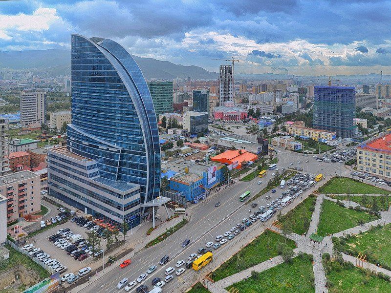 Cityscape with a large, curved blue glass skyscraper dominating the view, in Ulaanbaatar, Mongolia, with mountains in the background.