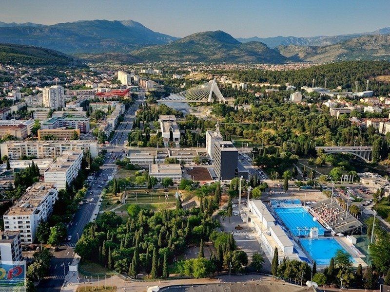 Aerial view of a city with a large pool, trees, buildings, a bridge, and mountains in the background.