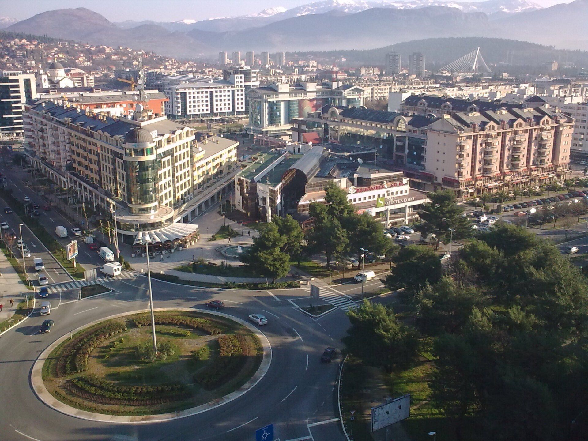Cityscape with buildings, a roundabout, and a distant mountain range under a sunny sky.