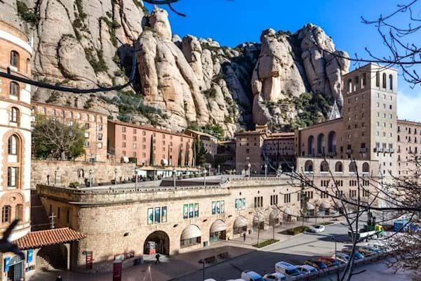 View of Montserrat Monastery, Spain, nestled at the base of a rugged mountain range, featuring a mix of architectural styles in tan and brown hues.