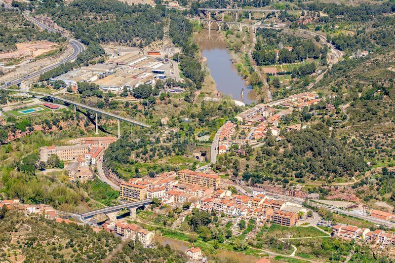 Aerial view of a town nestled in a valley with roads, bridges, and a river. The buildings have terracotta roofs, surrounded by green trees.