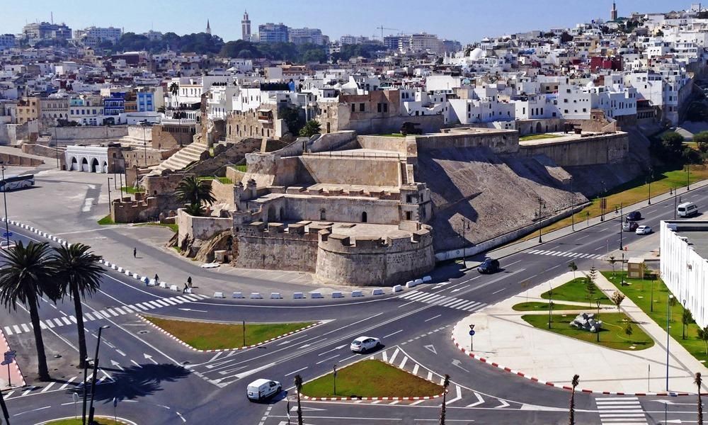 View of Tangier, Morocco. A fortress sits beside a road intersection with a city backdrop. Palm trees are in the foreground.