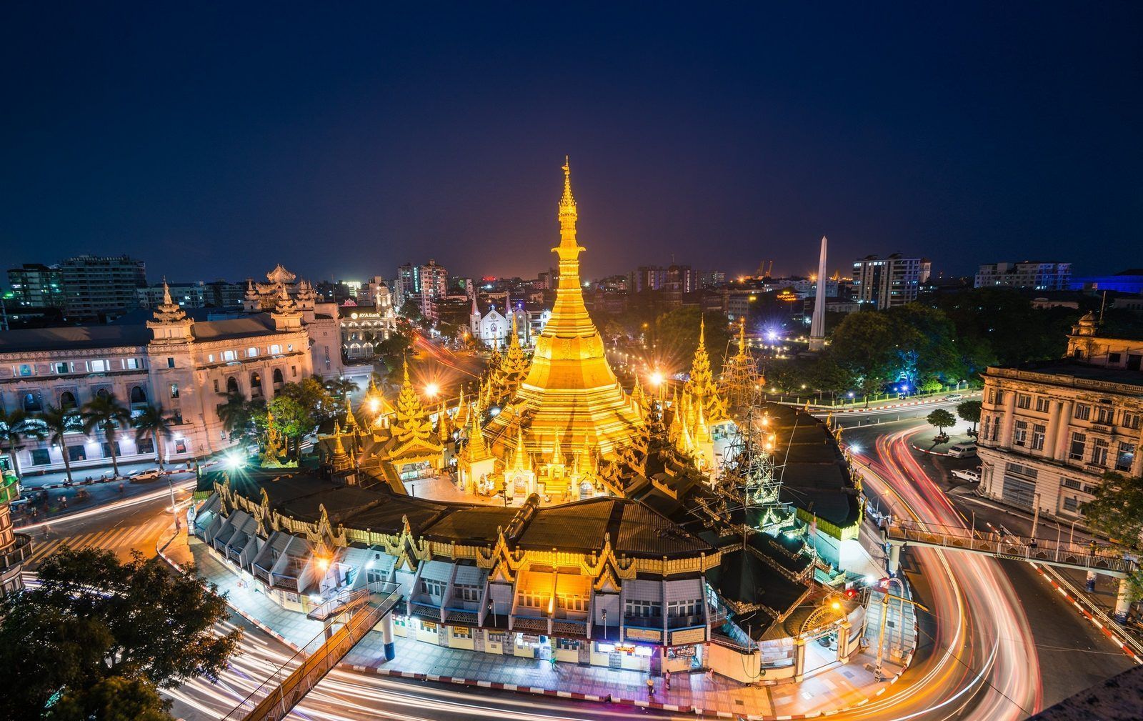 Golden Shwedagon Pagoda at night in Yangon, Myanmar, illuminated by lights, surrounded by cityscape and moving car lights.
