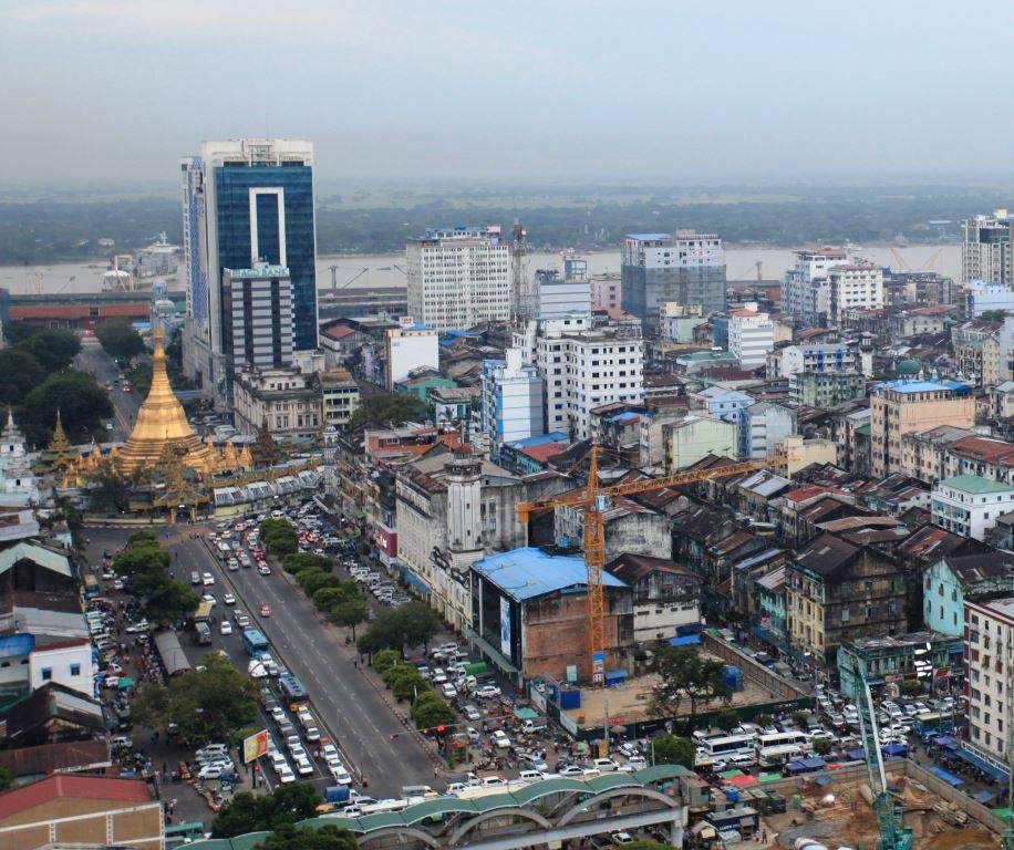 Aerial view of Yangon, Myanmar, featuring the golden Sule Pagoda, modern skyscrapers, and bustling city streets with dense traffic under a cloudy sky.