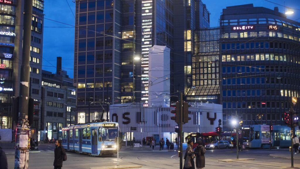 A tram travels through a city square at dusk, surrounded by tall buildings with lit windows. People stand on the street.