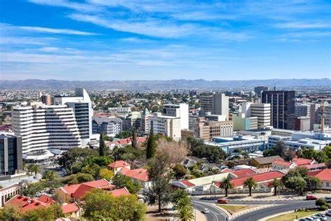 Cityscape of Windhoek, Namibia, under a blue sky. Modern buildings and red-roofed structures are visible.