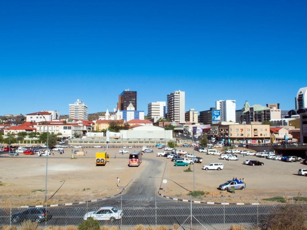 Skyline of Windhoek, Namibia, with buildings of varying heights against a clear blue sky. A large parking lot is in the foreground, with cars.