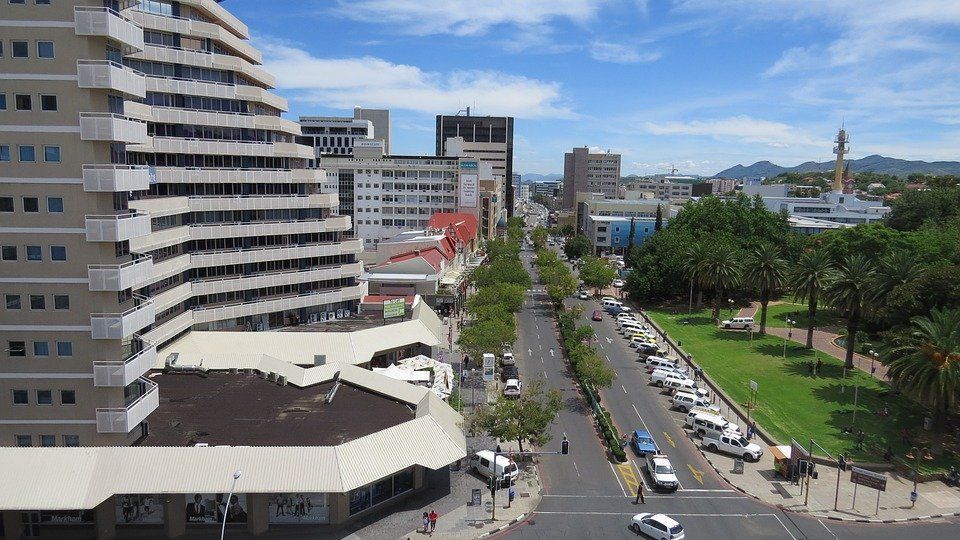 City street view in Windhoek, Namibia, with a tall building on the left, a tree-lined avenue, and a park on the right under a blue sky.