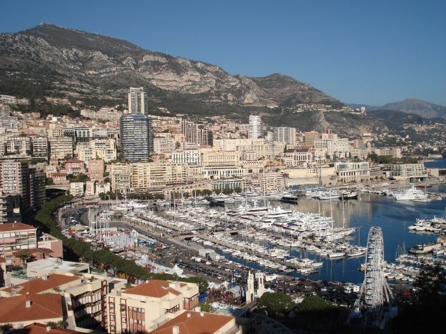 View of Monaco's harbor, filled with yachts and surrounded by buildings and mountains under a sunny sky.