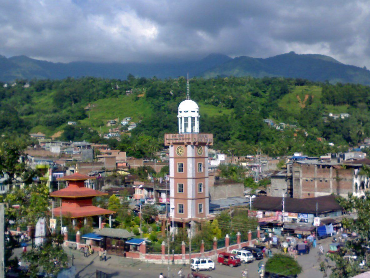 Clock tower in a town square with buildings, people, and mountains in the background under a cloudy sky.