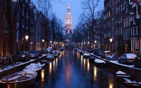 A canal in Amsterdam at dusk, with snow on the boats and buildings lining the water. A church tower rises in the distance.