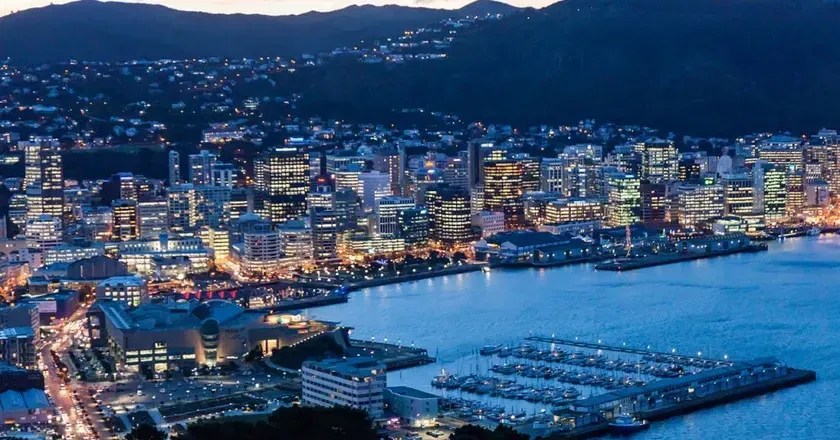 A city skyline at dusk, Wellington, New Zealand, with lights reflecting on the water. Buildings are lit along the coast.
