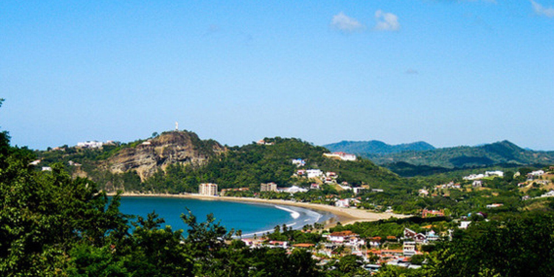 Scenic view of a beach nestled between green hills under a blue sky. Buildings dot the landscape, with a large rock formation on the left.
