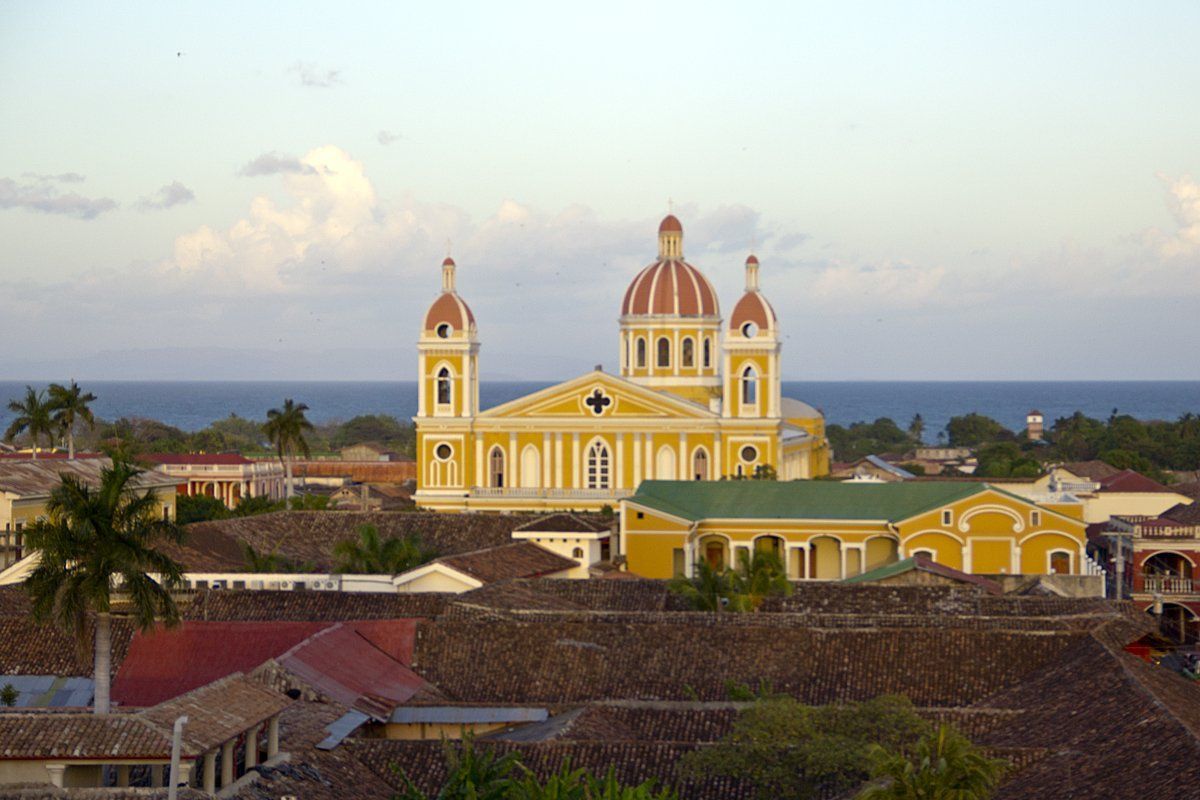 Yellow cathedral with red-domed towers and surrounding rooftops in Granada, Nicaragua, with the ocean visible in the background.