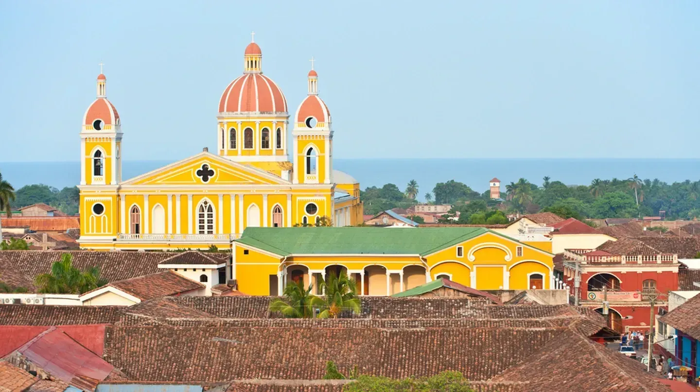 Yellow cathedral with red-topped domes overlooking a town with red-tiled roofs, near a body of water under a blue sky in Granada, Nicaragua.