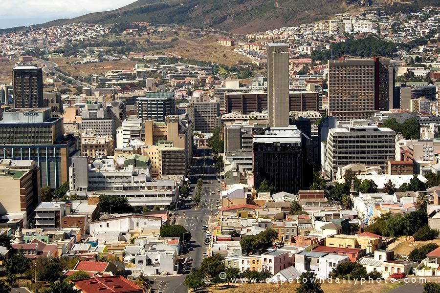 Cityscape of Cape Town, South Africa, featuring buildings of varying heights, a mountain in the background, and a wide road.