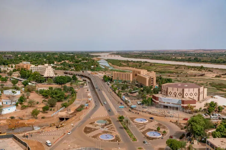 Aerial view of Niamey, Niger, showing a wide road with traffic, roundabouts, and buildings with beige and brown coloring. A river flows in the background.