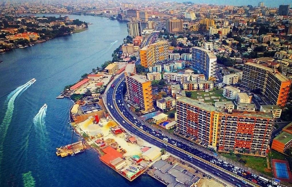 Aerial view of Lagos, Nigeria, with a busy waterfront and modern buildings lining the coast. A wide waterway divides the city.
