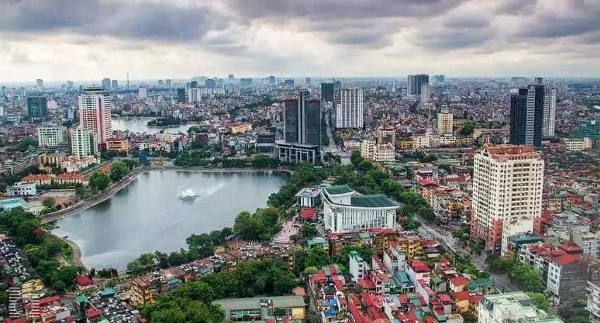 Aerial view of Hanoi, Vietnam, with a large lake in the foreground, surrounded by trees and buildings. The cityscape stretches into the distance under a cloudy sky.