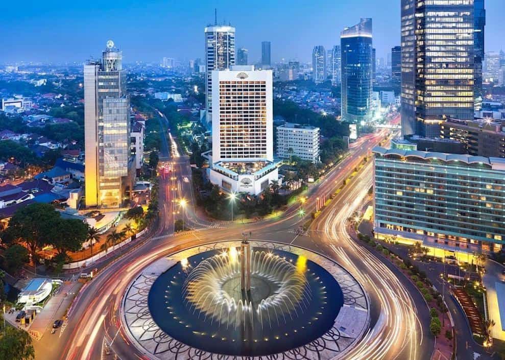 Jakarta cityscape at dusk, with a large roundabout featuring a fountain and busy traffic trails. Modern skyscrapers dominate the skyline.