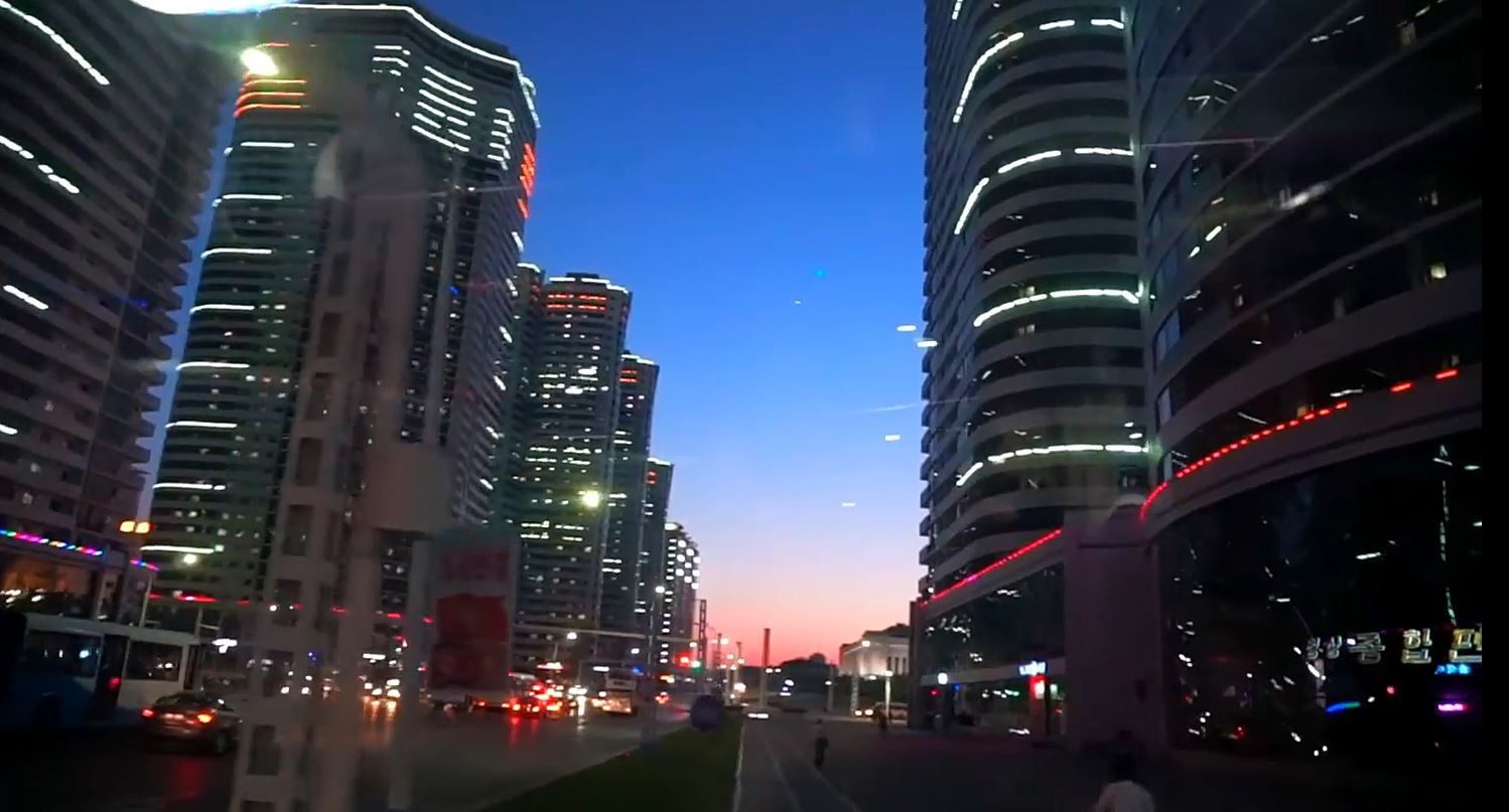 City skyline at dusk with tall buildings illuminated against a blue and pink sky, streetlights and traffic visible below.