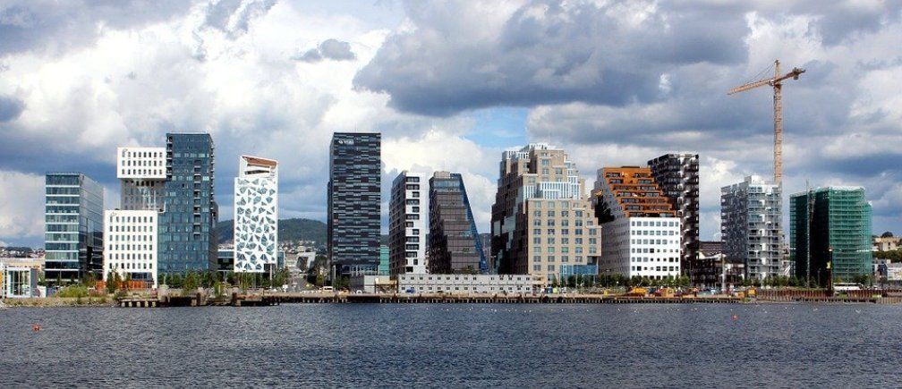 Skyline of Oslo, Norway, with modern architecture lining the waterfront under a cloudy sky. The sea is in the foreground.