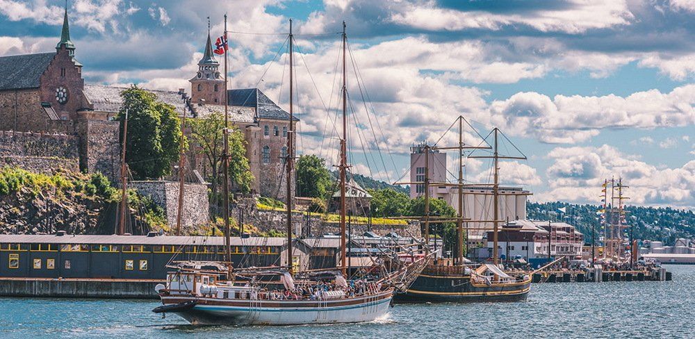 Sailboats on a harbor with historic buildings in the background under a cloudy sky.