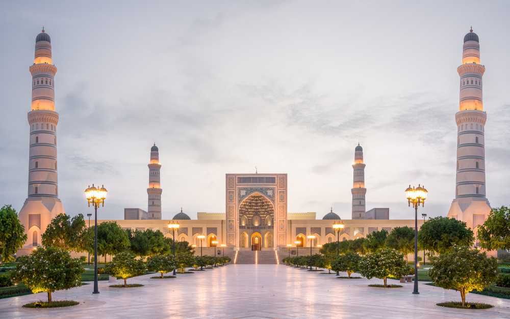 A grand mosque with towering minarets in Oman. Beige building with lit lights and a long, tree-lined walkway.