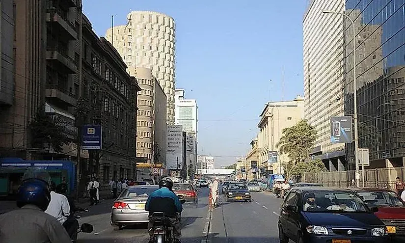 Street scene in Karachi, Pakistan, with cars, motorcycles, pedestrians, and tall buildings under a bright blue sky.