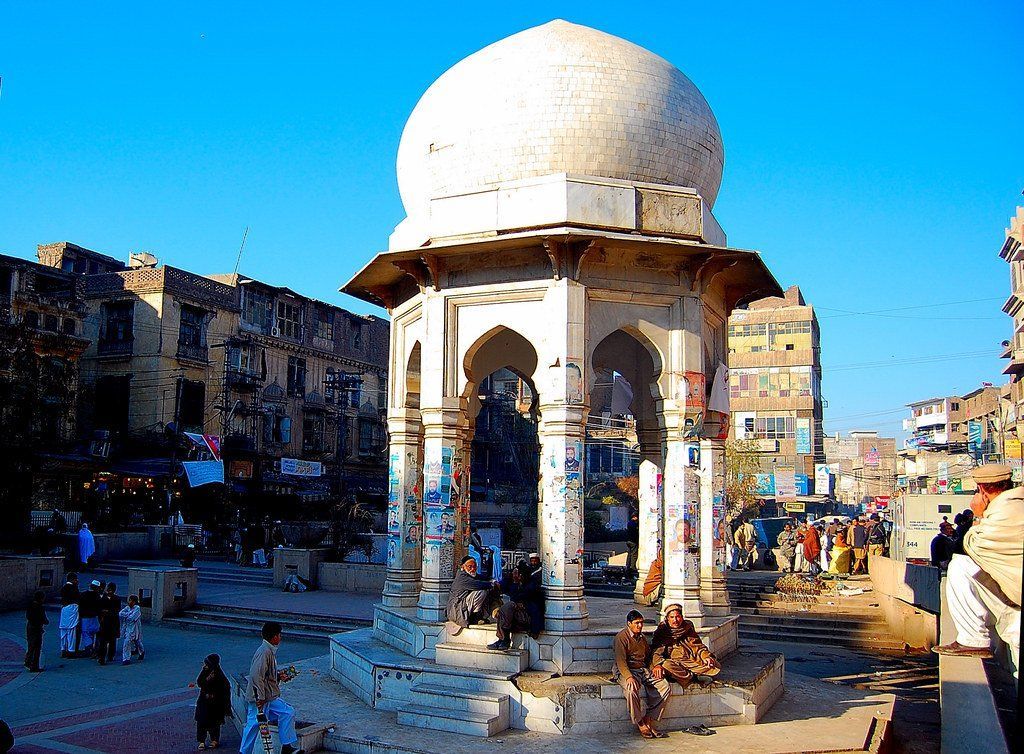 White domed structure in a bustling Pakistani market. People in traditional clothing gather near and around the ornate monument.