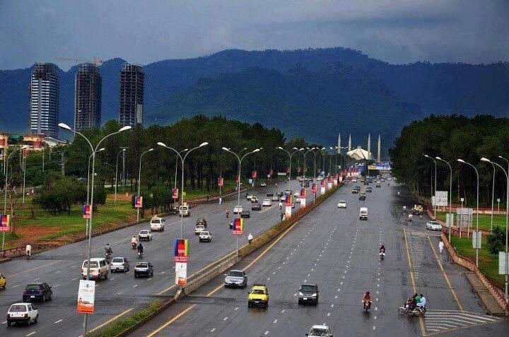 A wide road in Islamabad, Pakistan, with cars and bikes, flanked by trees and adorned with banners, with mountains in the background.