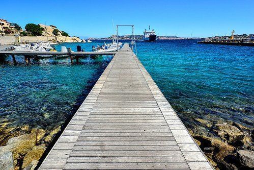 Wooden pier extending into turquoise water under a clear blue sky, boats docked on one side, distant ships and buildings.