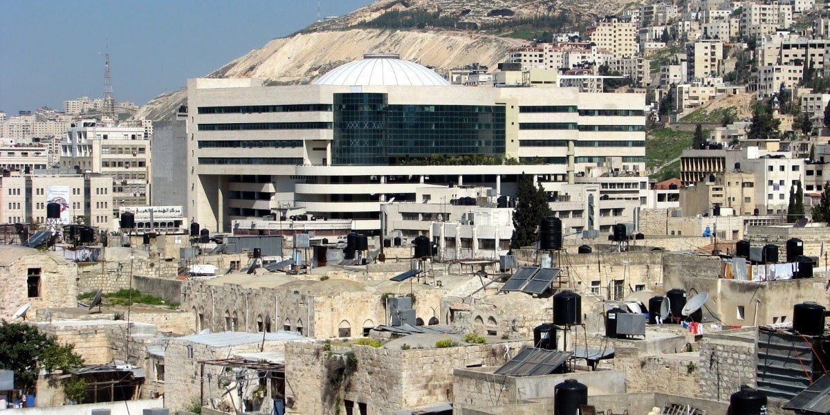 A large modern building with a domed roof dominates the skyline in a densely built city, seen over the rooftops of older buildings.