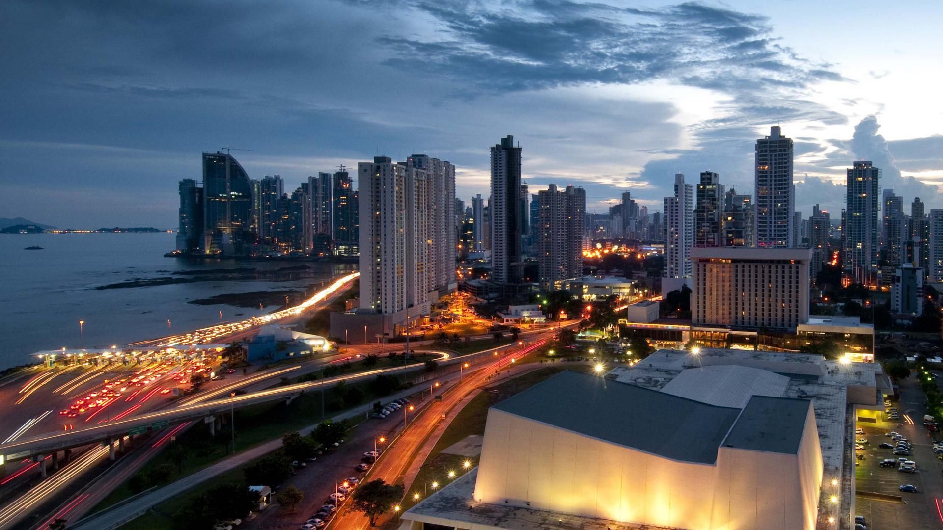 Nighttime cityscape of Panama City with illuminated skyscrapers, roads, and coastline under a twilight sky.