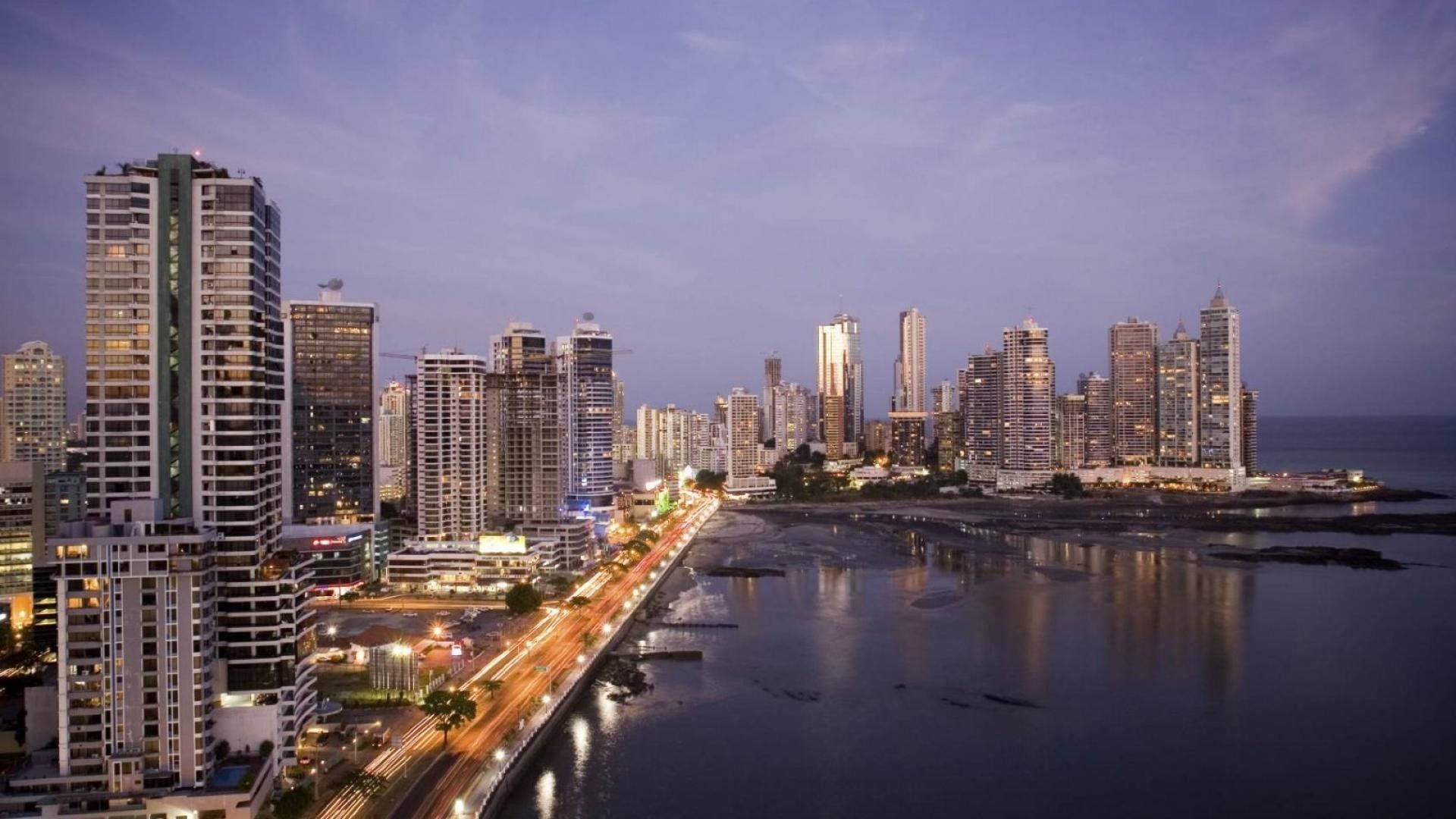 City skyline at dusk with illuminated skyscrapers along a coastal road. Lights reflect on the water.