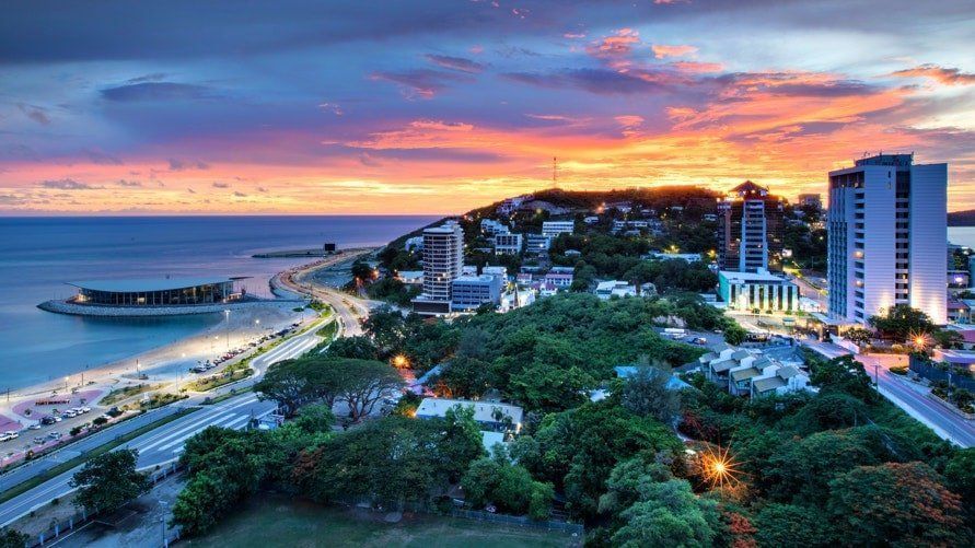 Coastal city at sunset: Buildings and a road line the shore beneath a colorful sky.