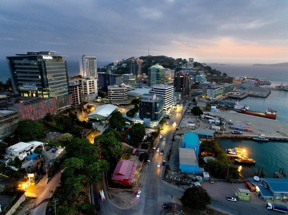 An aerial view of Port Moresby, Papua New Guinea, at dusk. Buildings line the coast, with a harbor and hill in the background.