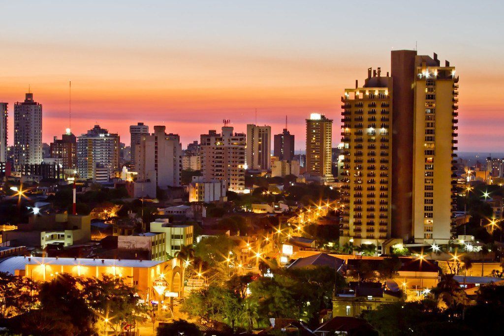 City skyline at sunset, with illuminated buildings and streetlights under a colorful orange and pink sky.
