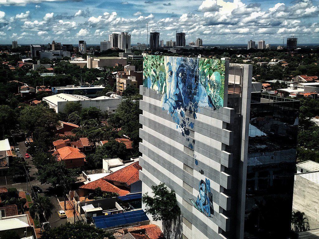 Cityscape with a tall modern building featuring a colorful mural. Buildings and trees fill the view under a cloudy sky.