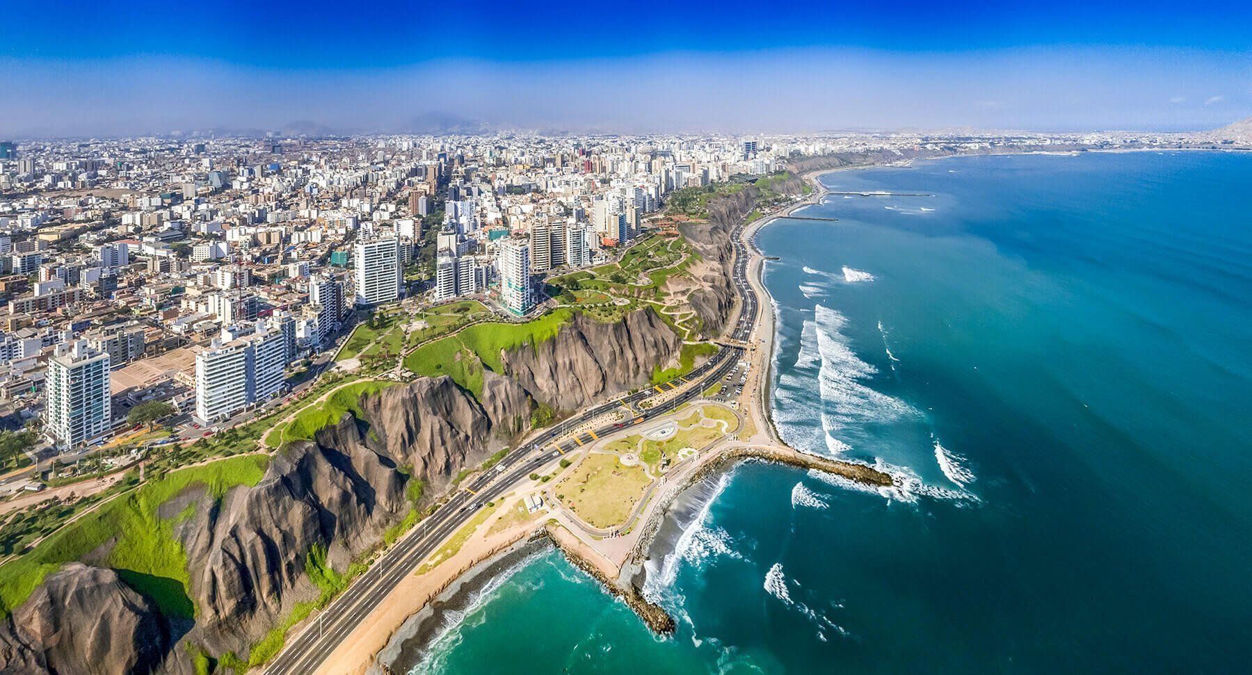 Aerial view of Lima, Peru, with cityscape meeting the Pacific Ocean. Turquoise water meets a rocky cliffside, with a coastal road and buildings.