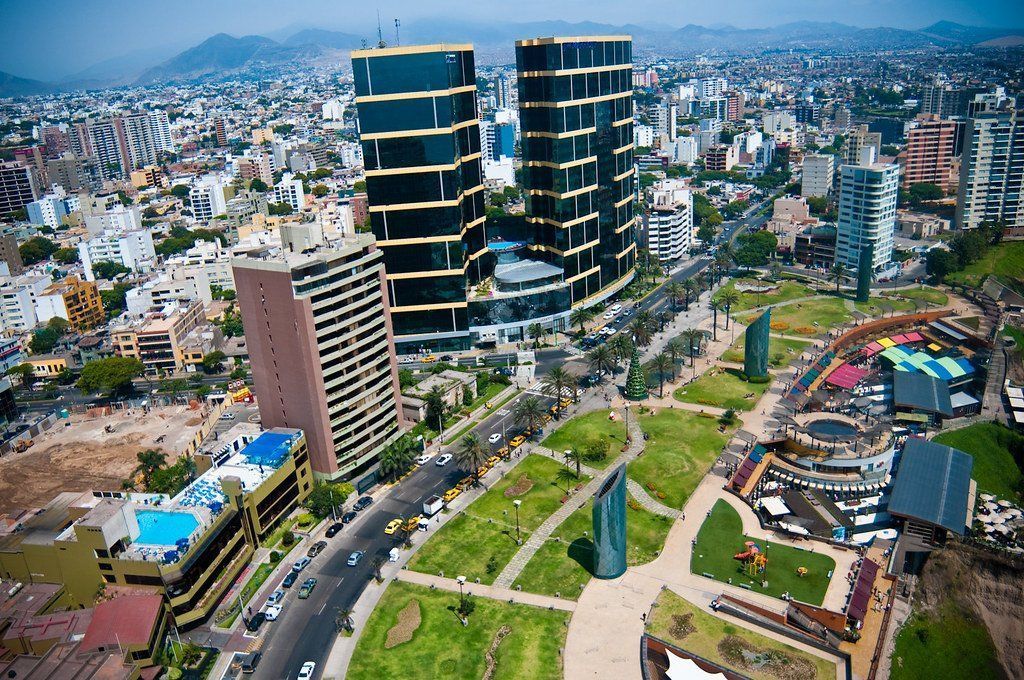 Aerial view of Miraflores, Lima, Peru, with modern skyscrapers overlooking a park and city. Blue skies and mountain backdrop.
