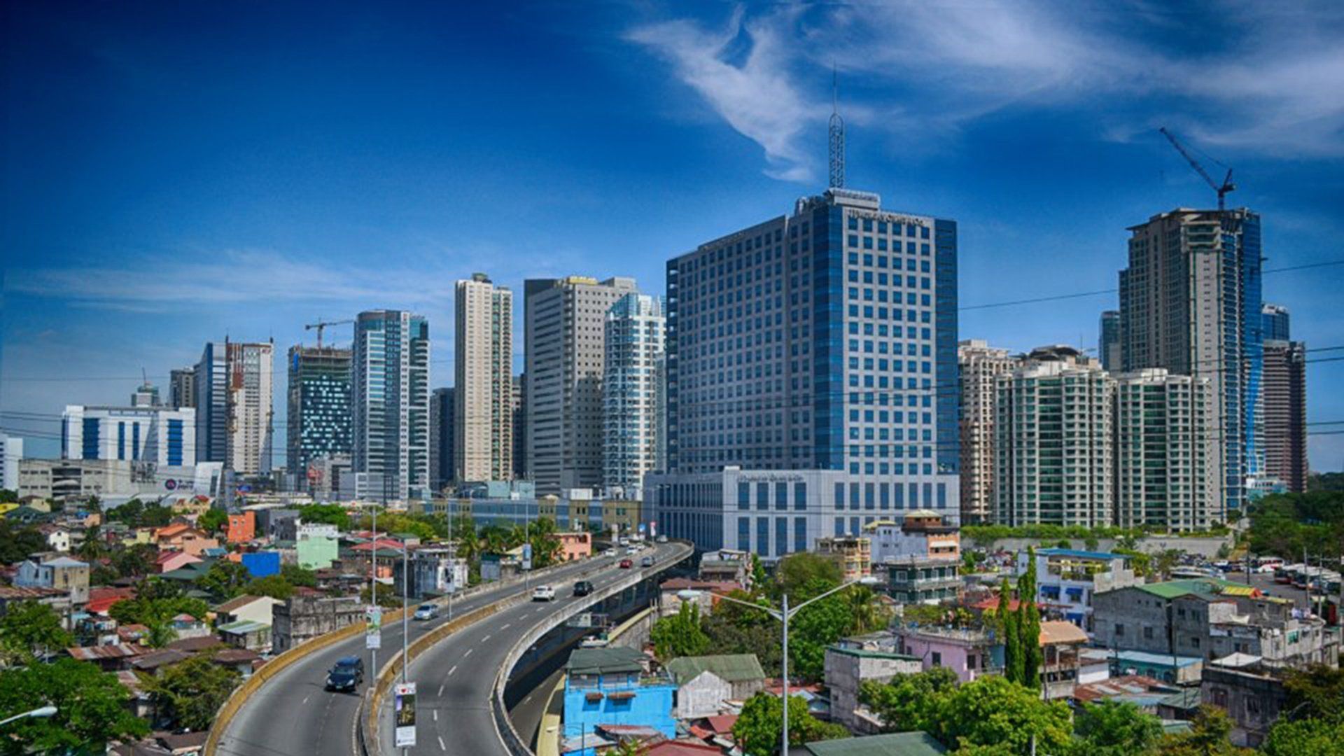 Skyline of a city, with a mix of high-rise buildings and low-rise residential areas, under a blue sky. A highway runs through the foreground.