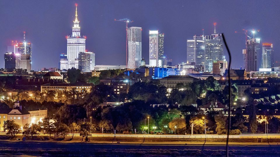 Nighttime cityscape with illuminated skyscrapers and buildings under a dark blue sky, likely Warsaw, Poland.