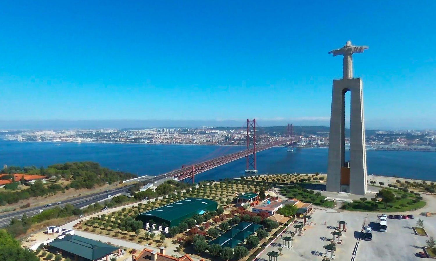 Christ the King statue overlooking Lisbon, Portugal, with the 25 de Abril Bridge and city skyline visible. Clear blue sky.