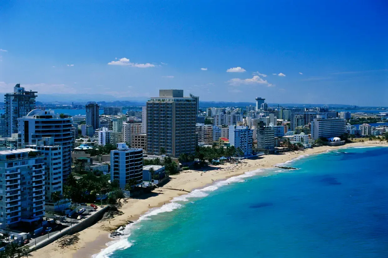 Coastal city skyline with a sandy beach and bright blue water under a clear sky.